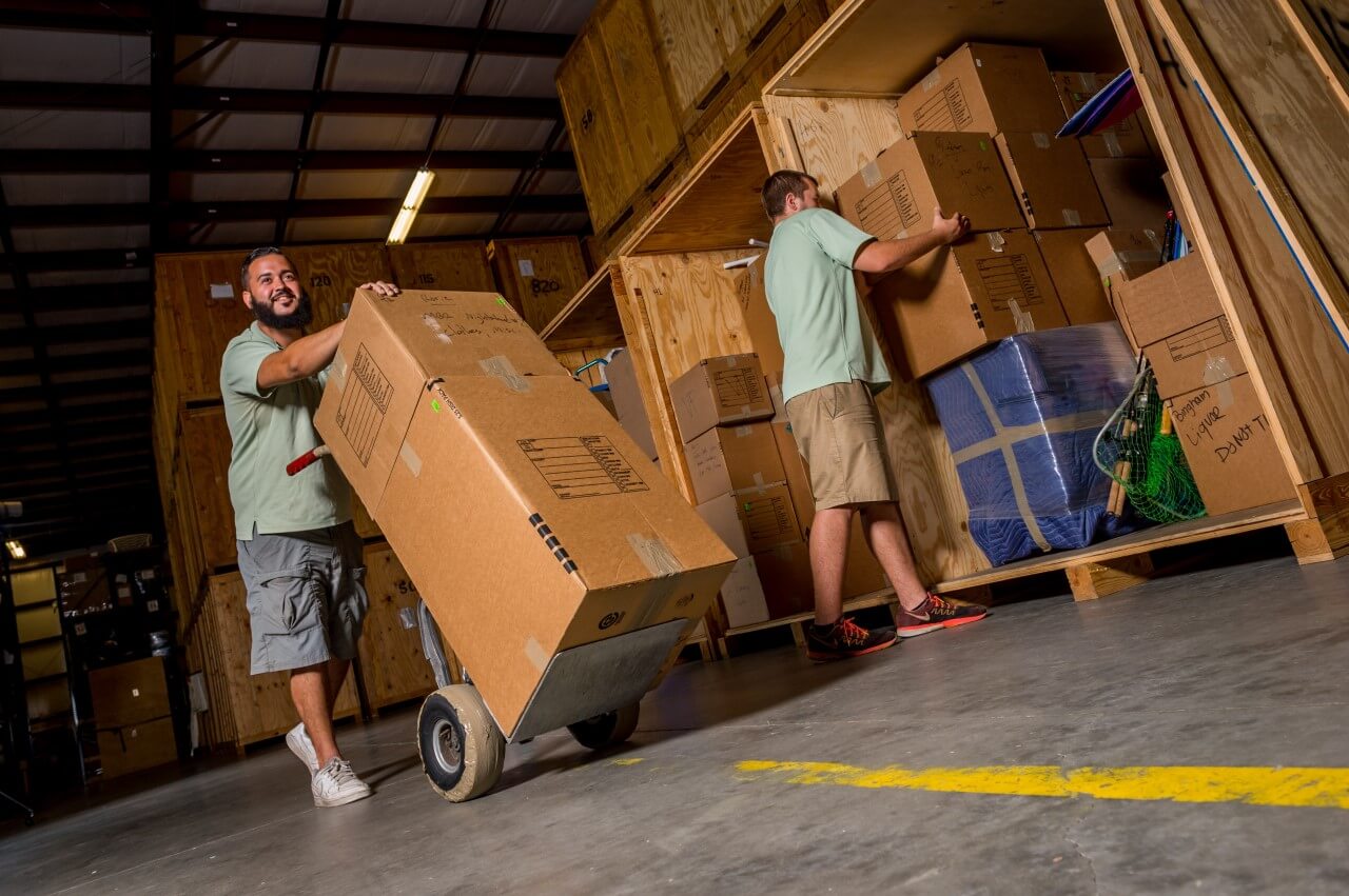 Professional movers using a hand truck to load boxes into a wooden storage vault in a warehouse