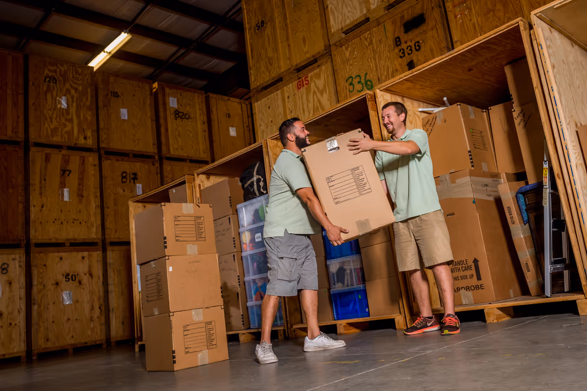 Two movers in matching uniforms lifting a large cardboard box into a wooden storage vault
